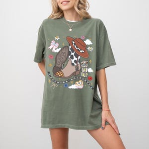 May include: An olive green short-sleeve t-shirt featuring a graphic print of a brown cowboy hat, a cow-print western boot with the number 13, alphabet blocks spelling TS, a beaded necklace, a pink bow, flowers, and clouds on the front center.