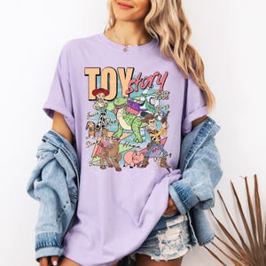May include: A light purple short-sleeve t-shirt featuring a graphic print of Toy Story characters including Woody, Buzz Lightyear, Jessie, Rex, Slinky Dog, Bullseye, and Hamm with the text Toy Story Since 1995 and individual character names.