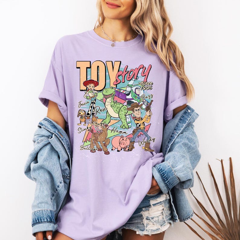May include: A light purple short-sleeve t-shirt featuring a graphic print of Toy Story characters including Woody, Buzz Lightyear, Jessie, Rex, Slinky Dog, Bullseye, and Hamm with the text Toy Story Since 1995 and individual character names.