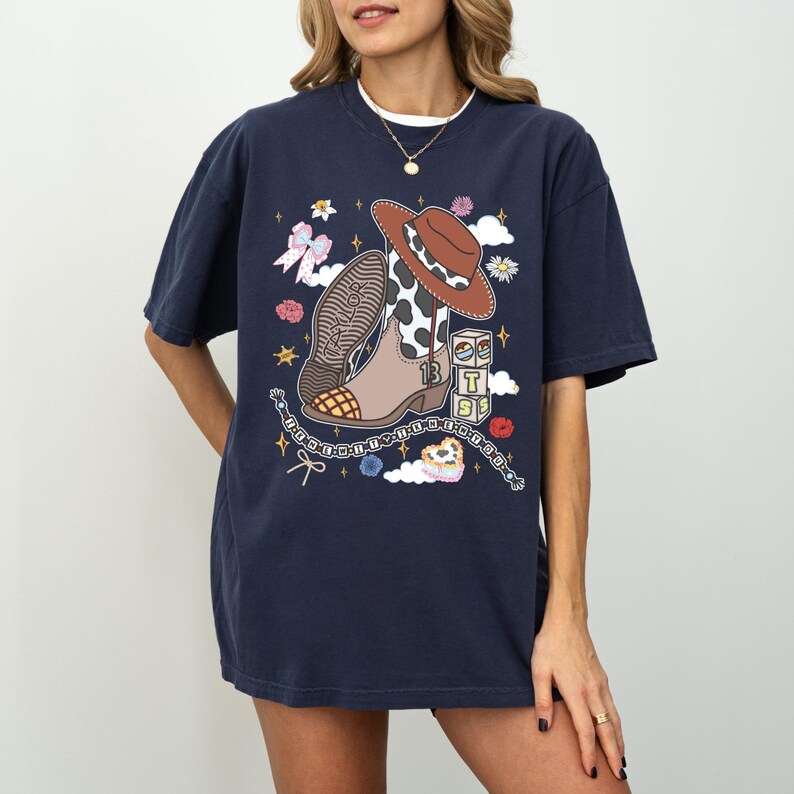 May include: A navy blue short-sleeve t-shirt featuring a graphic print of a brown and white cow-print cowboy boot, a brown hat, a stack of blocks with the letters T, S, and S, a pink bow, and various small star and flower illustrations.