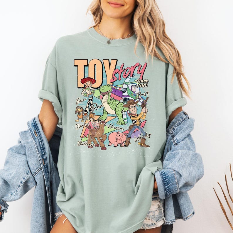 May include: A light sage green short-sleeve t-shirt featuring a colorful graphic print of Toy Story characters including Woody, Buzz Lightyear, Jessie, Rex, Hamm, Slinky Dog, and Bullseye with the text Toy Story Since 1995 and character names.