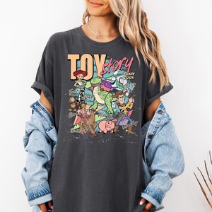 May include: A charcoal gray t-shirt featuring a graphic print of Toy Story characters including Woody, Buzz Lightyear, Jessie, Rex, Slinky Dog, and Hamm. The text reads TOY story Since 1995, with character names written in cursive script.