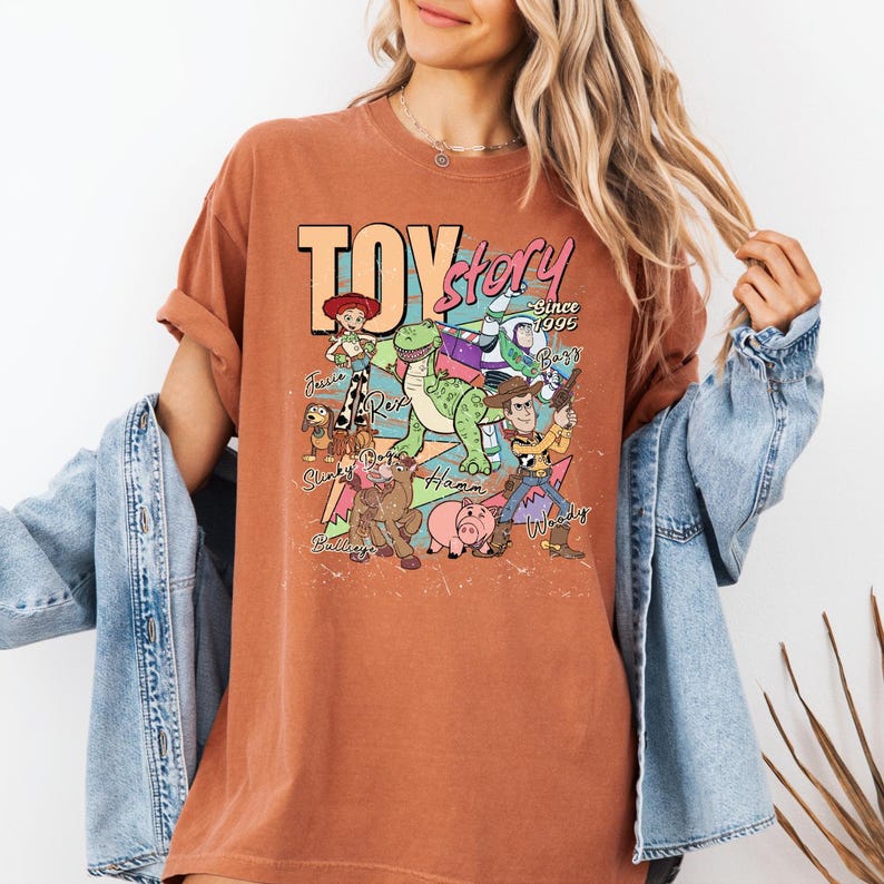 May include: A rust-colored short-sleeve t-shirt featuring a graphic print of Toy Story characters including Woody, Buzz Lightyear, Jessie, Rex, Slinky Dog, Bullseye, and Hamm. The text reads Toy Story Since 1995 with character names printed below.