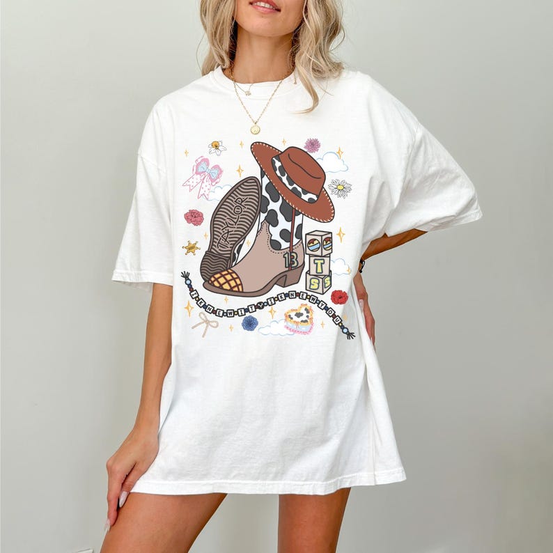May include: A white short-sleeve t-shirt featuring a graphic print of a brown and white cow-print cowboy boot, a brown cowboy hat, a beaded bracelet, and stacked blocks with the letters T, S, and the number 13, surrounded by small floral icons.