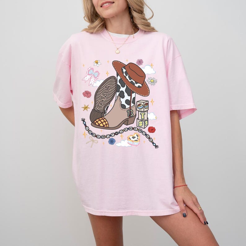 May include: A light pink t-shirt featuring a graphic print of a brown and white cow-print cowboy boot, a brown cowboy hat, and three stacked blocks with the letters T, S, and S. The design includes a beaded string with the text TAYLOR SWIFT.