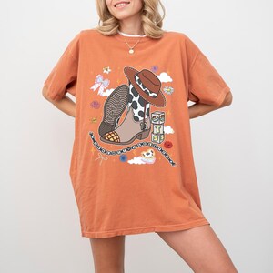 May include: An orange short-sleeve t-shirt featuring a graphic print of a brown and white cow-print cowboy boot, a brown hat, three stacked blocks with the letters T, S, and a heart, a beaded bracelet, a bow, and small floral accents.