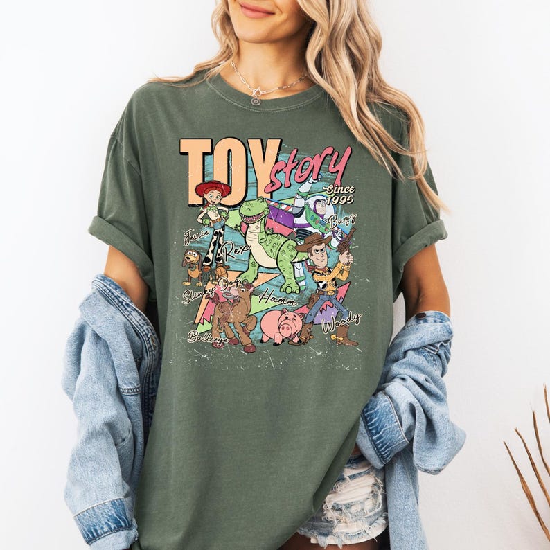 May include: An olive green short-sleeve t-shirt featuring a graphic print of Toy Story characters including Woody, Buzz Lightyear, Jessie, Rex, Slinky Dog, Bullseye, and Hamm. The text reads Toy Story Since 1995 with character names printed in script.