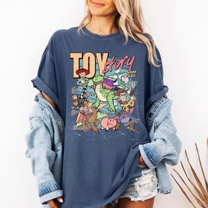 May include: A dark blue t-shirt featuring a graphic print of Toy Story characters including Woody, Buzz Lightyear, Jessie, Rex, Slinky Dog, Hamm, and Bullseye. The text reads Toy Story Since 1995 with character names written in cursive script.