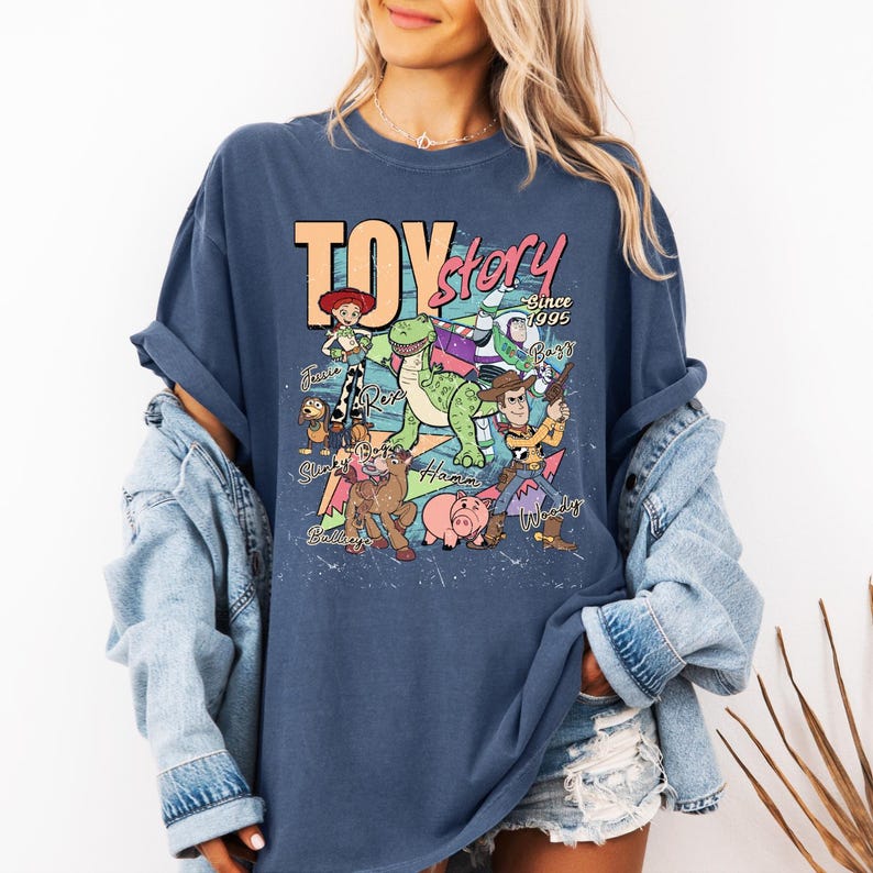 May include: A dark blue t-shirt featuring a graphic print of Toy Story characters including Woody, Buzz Lightyear, Jessie, Rex, Slinky Dog, Hamm, and Bullseye. The text reads Toy Story Since 1995 with character names written in cursive script.