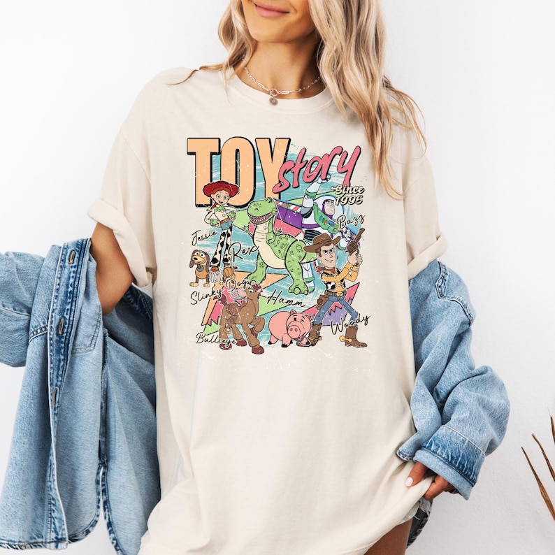 May include: A cream-colored short-sleeve t-shirt featuring a graphic print of Toy Story characters including Woody, Buzz Lightyear, Jessie, Rex, Slinky Dog, Bullseye, and Hamm with the text Toy Story Since 1995 and individual character names.