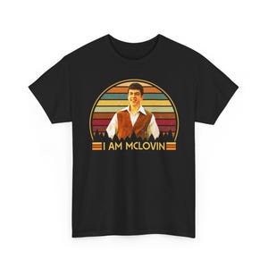 May include: Black t-shirt featuring a retro design with a man in a brown vest and white shirt, set against a sunset backdrop. The text "I AM MCLOVIN" is displayed below the image in a vintage style.