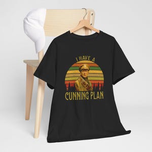 May include: Black t-shirt with a graphic featuring a man in a military uniform, set against a sunset design with the text "I HAVE A CUNNING PLAN". A white baseball cap rests on a wooden chair.