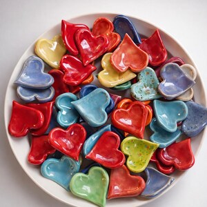 May include: A white ceramic bowl filled with colorful, heart-shaped ceramic dishes. The hearts are in shades of red, blue, yellow, and green. The glossy finish of the hearts contrasts with the matte white of the bowl.