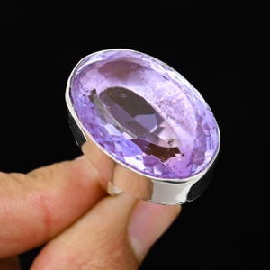 Pode incluir: Anel de prata com uma grande ametista oval. A ametista tem um tom roxo claro e é definida em uma moldura de prata polida. O anel é mostrado em um fundo preto.