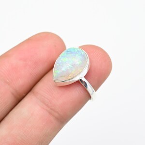 May include: A silver ring featuring a pear-shaped opal gemstone with iridescent flashes of green, blue, and yellow. The stone is set in a smooth metal bezel, and the band is a simple, polished silver wire that encircles the finger.