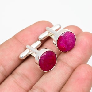 May include: A pair of silver-toned cufflinks featuring round, faceted, deep pinkish-red gemstone cabochons. Each cufflink has a polished metal toggle backing with a cylindrical post and a hinged bar mechanism for securing through shirt buttonholes.