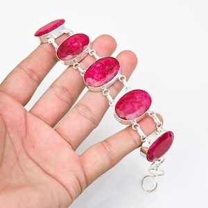 May include: A silver bracelet featuring five large, oval-shaped, faceted deep pink ruby gemstones set in polished metal bezels. The stones are connected by silver links, and the bracelet includes a small circular clasp at the end of the chain.
