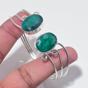 May include: A silver cuff bracelet featuring two oval-shaped, faceted green gemstones. The metal band consists of three thin, parallel silver wires that curve into decorative loops at the points where they connect to the gemstone settings.