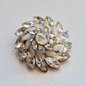 May include: A circular brooch featuring a central round cabochon surrounded by alternating layers of marquise-cut clear crystals and smooth, opaque white oval stones arranged in a radiating, floral-inspired pattern with a metallic base.