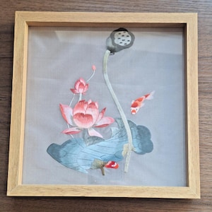 May include: Embroidered artwork of a lotus flower with pink petals, a gray seed head, and a blue-green leaf. Two koi fish swim near the flower. The artwork is framed in a light brown wooden frame.