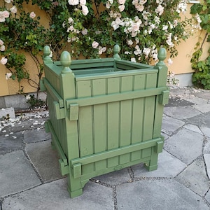 Puede incluir: Una jardinera cuadrada de madera color verde salvia con listones verticales, vigas de soporte horizontales y cuatro remates decorativos en cada poste de esquina. La estructura está construida con un marco resistente y se apoya sobre cuatro patas.