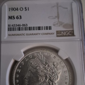 May include: A silver 1904 O $1 Morgan dollar coin, graded MS 63, in a protective holder. The holder displays "1904 O $1", "MS 63", "8143346-063", and "NUMISMATIC GUARANTY COMPANY" with the NGC logo. The coin's obverse is visible, showing the inscription.