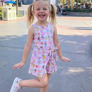 Może przedstawiać: Małe dziecko w r&oacute;żowym romperze z wzorem Myszki Miki i serduszek oraz pasujących butach. Tło przedstawia zamek, karuzelę i palmy, sugerując park rozrywki.