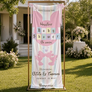 May include: A baby shower banner with a pastel rainbow striped background. The banner features the text "Happiest Baby Shower On Earth" and "Honoring Olivia & Thomas". Two pink silhouettes of Mickey Mouse are at the bottom. The banner is displayed on a gold frame.