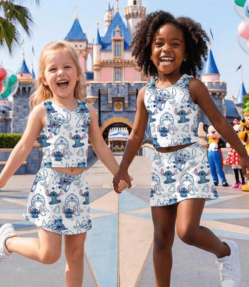 May include: Two young girls wearing matching white crop tops and skirts with a blue Stitch character pattern. They are holding hands and smiling in front of a castle, with balloons and cartoon characters in the background.