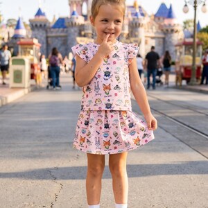 Puede incluir: Una ni&ntilde;a peque&ntilde;a con un conjunto rosa de dos piezas con estampado de personajes de dibujos animados. El conjunto incluye una camiseta con mangas con volantes y una falda plisada. El fondo muestra un castillo y un d&iacute;a soleado.