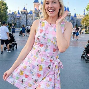 Może przedstawiać: R&oacute;żowo-biały kombinezon w kratę z postaciami Disneya i balonami. Bezrękawnik ma sp&oacute;dnicę kopertową i jest wiązany w talii. Tło obejmuje zamek i ludzi.