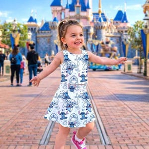 May include: A sleeveless white dress and skirt set featuring a repeating pattern of a blue cartoon character. The outfit is worn with pink sneakers. The background shows a large castle and a sunny day.