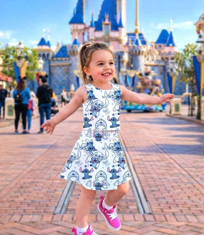 May include: A sleeveless white dress and skirt set featuring a repeating pattern of a blue cartoon character. The outfit is worn with pink sneakers. The background shows a large castle and a sunny day.