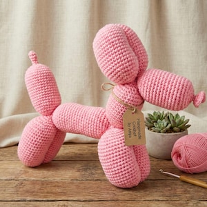 Puede incluir: Un perro globo rosa hecho a mano con ganchillo. El perro está hecho de hilo suave y tiene una etiqueta que dice "Handmade Crochet by Anya". También se ven una madeja de hilo rosa, un ganchillo y una pequeña suculenta en maceta.