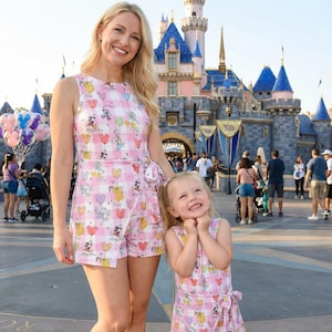 Może przedstawiać: Kobieta i małe dziecko noszą pasujące r&oacute;żowo-białe rompers w kratkę z grafiką balon&oacute;w Myszki Miki. Stoją przed bajkowym zamkiem z niebieskimi wieżami. Stroje mają prz&oacute;d w stylu kopertowym i kokardę w talii.