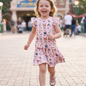 Puede incluir: Una ni&ntilde;a peque&ntilde;a con un conjunto rosa de dos piezas con un estampado de animales de dibujos animados, incluyendo zorros y conejos, corre hacia la c&aacute;mara. El conjunto consta de una camiseta de manga corta con volantes y una falda a juego. La ni&ntilde;a lleva zapatillas rosas.