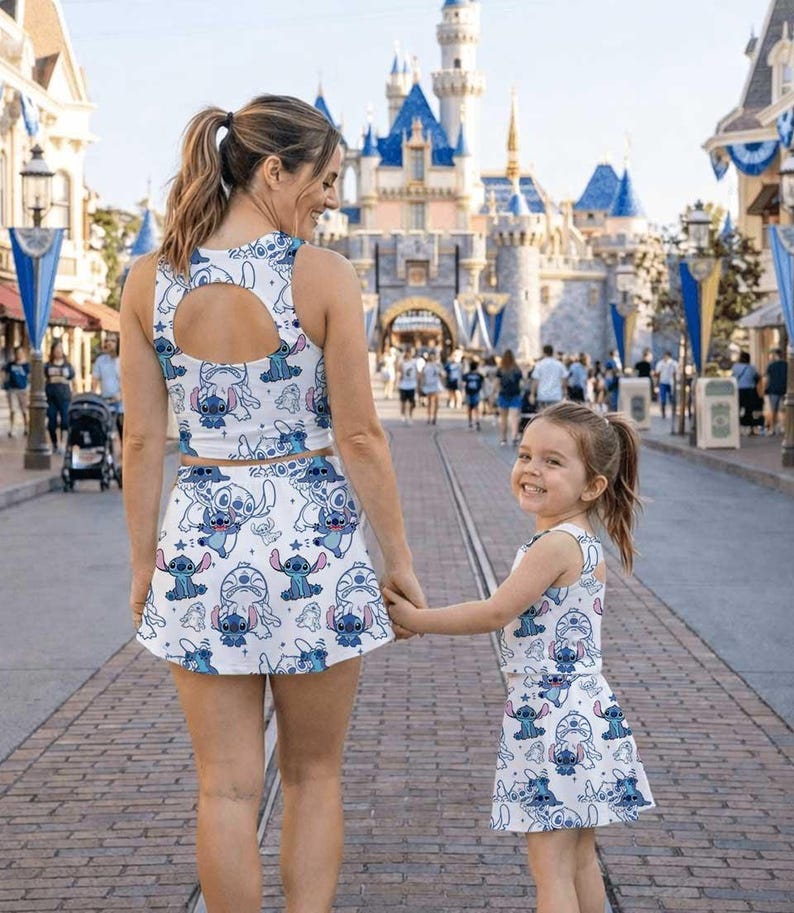 May include: Matching white and blue outfits featuring a cartoon character print. The outfits include crop tops and skirts. A woman and a child are holding hands and walking on a brick road in front of a castle.