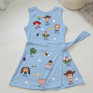 May include: A light blue sleeveless dress featuring a wrap-style skirt with a tie closure. The fabric is printed with various Toy Story character faces including Woody, Jessie, Bo Peep, Buzz Lightyear, Rex, Bullseye, Slinky, and Hamm with names.