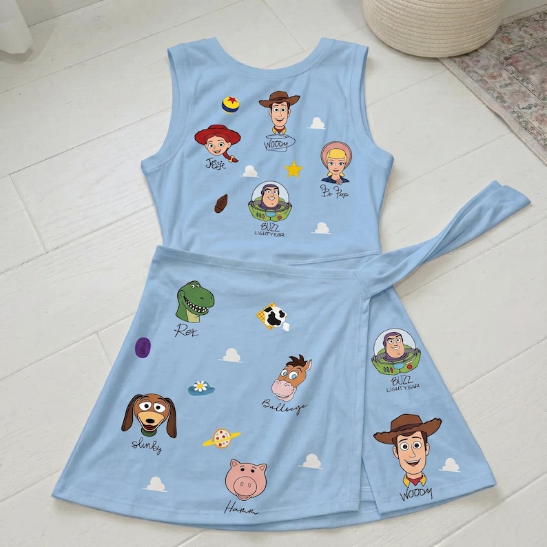 May include: A light blue sleeveless dress featuring a wrap-style skirt with a tie closure. The fabric is printed with various Toy Story character faces including Woody, Jessie, Bo Peep, Buzz Lightyear, Rex, Bullseye, Slinky, and Hamm with names.