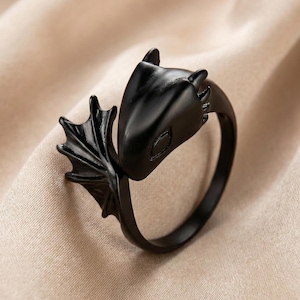 May include: A black dragon ring with a detailed head and wing design. The ring is made of a dark metal and features a smooth, polished finish. The dragon's head is facing forward, and the wings are spread out to the side. A unique piece of jewelry.