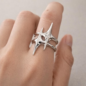 May include: A silver-colored ring with a unique, abstract design. The ring features a starburst-like shape with multiple pointed projections and a double-band design. The ring is worn on a finger, showcasing its modern and artistic style.