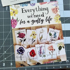 May include: A collage featuring floral illustrations and inspirational text. The main text reads "Everything we need for a godly life." Individual cards display words like "love," "joy," and "peace," each paired with a watercolor flower. The background is a cutting mat.