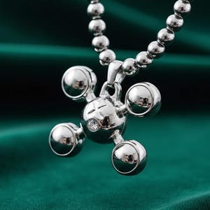 Puede incluir: Un collar con colgante plateado con un diseño esférico único. El colgante presenta una esfera central con cuatro esferas más pequeñas que se extienden hacia afuera, y una pequeña gema transparente. La cadena del collar está hecha de pequeñas cuentas redondas.