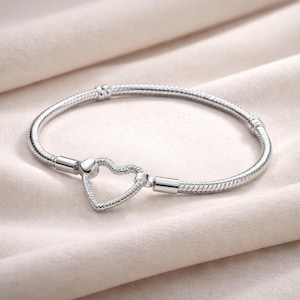 Pode incluir: Uma pulseira prateada com fecho em forma de coração. A pulseira tem um design de corrente de cobra e um fecho seguro. O fecho de coração está no centro da pulseira. A pulseira é exibida em uma superfície macia e clara.