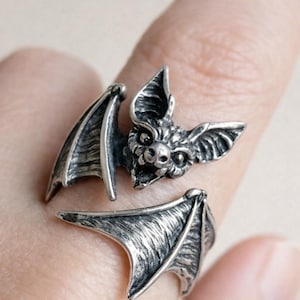 Puede incluir: Anillo de murciélago plateado con alas detalladas y superficie texturizada. La cabeza del murciélago está centrada, con las alas extendiéndose a ambos lados. El anillo envuelve un dedo, mostrando un tema gótico o de Halloween.