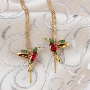 May include: Two gold-colored hummingbird necklaces. Each pendant features a red gemstone body, green gemstone accents, and gold wings. The necklaces hang on delicate gold chains, resting on a white, textured surface.