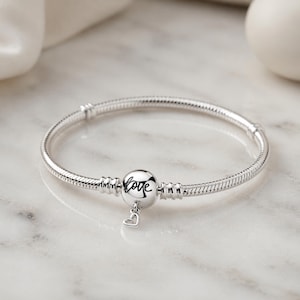 May include: A silver bracelet with a round charm that says "love" in cursive. A small heart charm dangles below the round charm. The bracelet has a snake chain design.