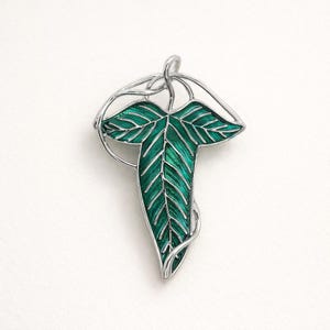 May include: A silver-toned leaf-shaped brooch with a vibrant green enamel fill. The leaf design features detailed veining and is surrounded by a delicate, intertwined silver wire frame. The brooch is set against a plain white background.