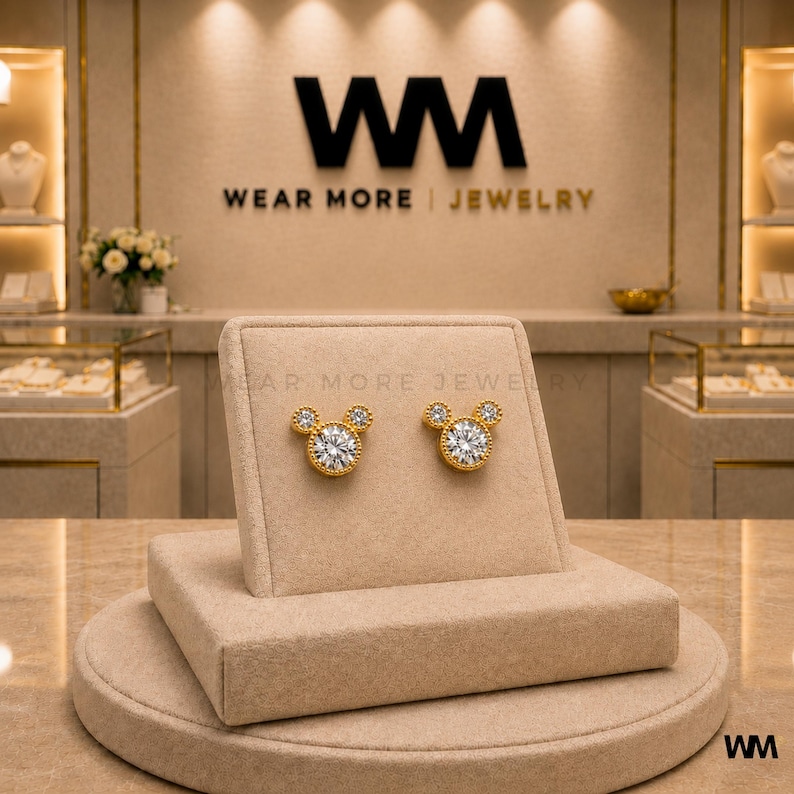 May include: A pair of gold-colored stud earrings in the shape of a mouse head, with a large, round, clear stone in the center and smaller stones for the ears. The earrings are displayed on a beige jewelry stand. The store's logo, "WM WEAR MORE JEWELRY," is visible in the background.