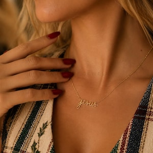 May include: A gold-toned necklace featuring a delicate script-style pendant that spells out the word "jesus" in lowercase cursive lettering. The thin, polished chain connects to the pendant on both sides, resting against the collarbone area.